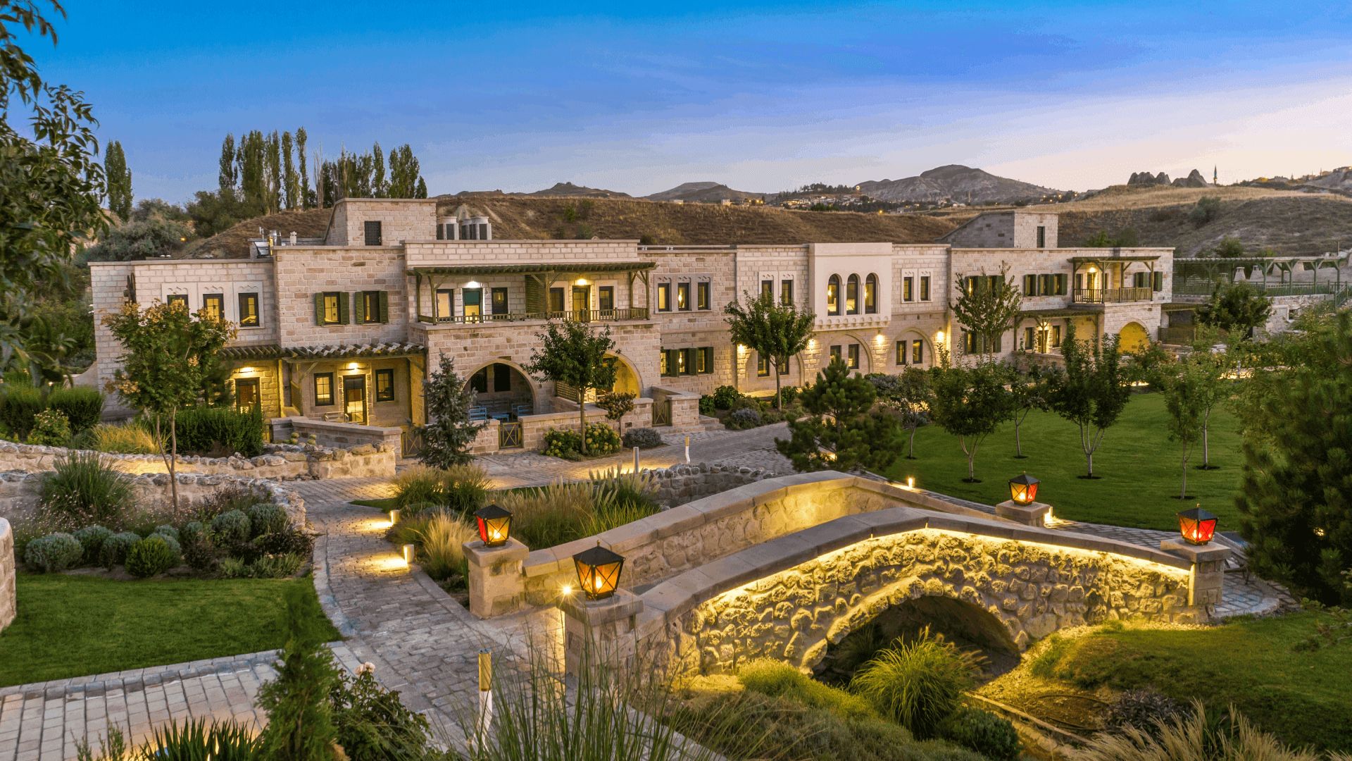 Complejo turístico de lujo con arquitectura de piedra, jardines y puente iluminado al atardecer.