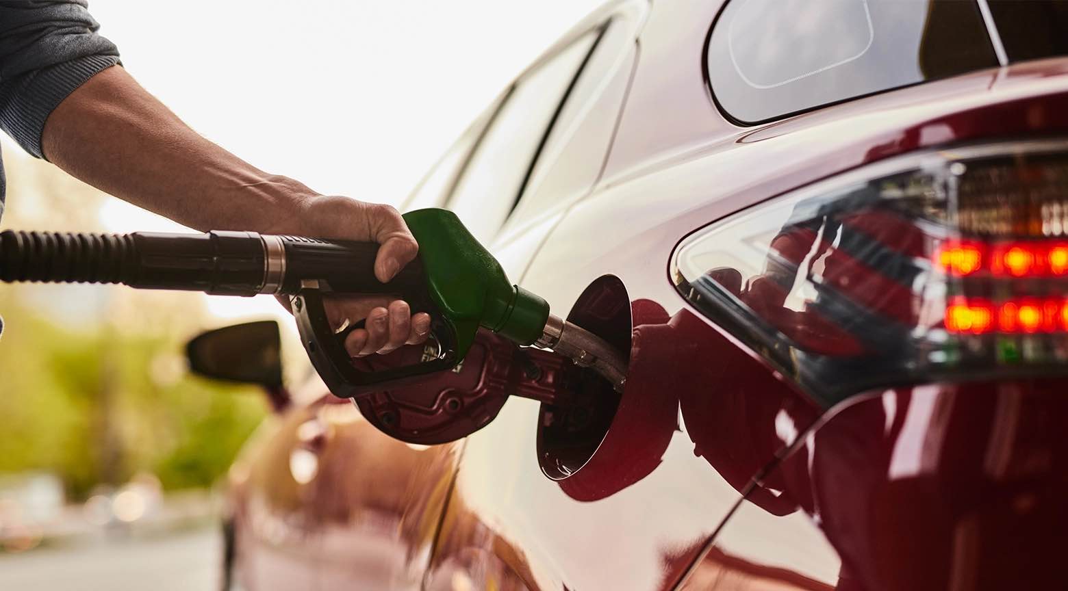 Primer plano de perfil del brazo y la mano de una persona que sostiene con firmeza una manguera de llenado con boquilla verde introducida en el tanque abierto de un automóvil de color rojo vino brillante del cual se llega a apreciar la parte trasera con el faro posterior encendido y el reflejo de la luz exterior sobre la carrocería limpia en una estación de servicios.