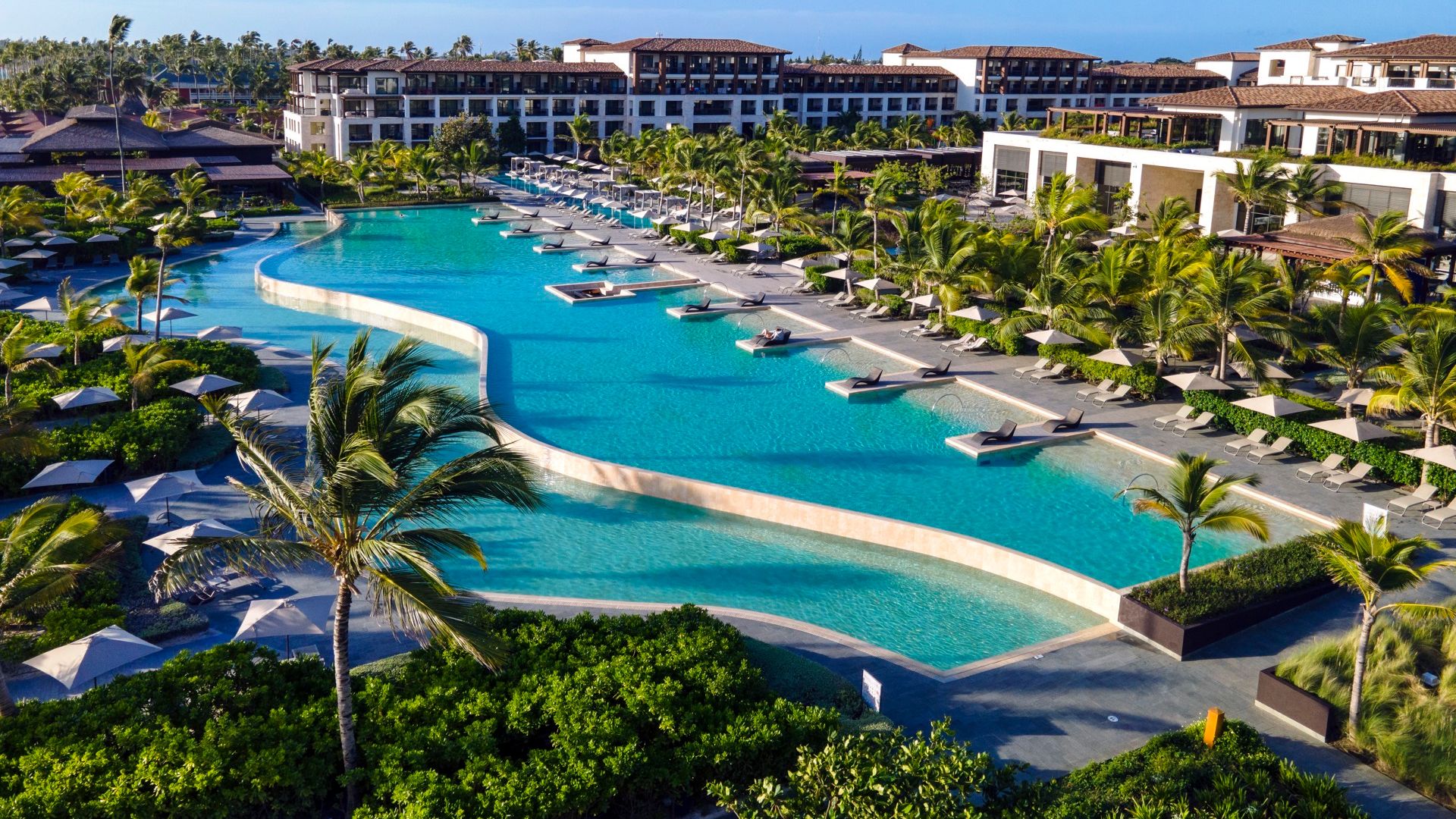 Vista aérea de una piscina de lujo en un resort en Punta Cana, rodeada de sombrillas y palmeras, con un diseño curvado.