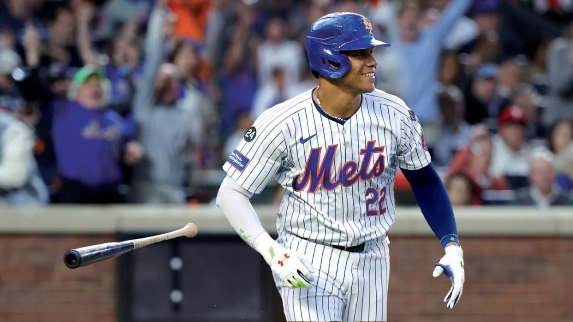 Jugador de béisbol de los New York Mets celebrando una jugada con el bate en el aire, mientras los aficionados lo animan.