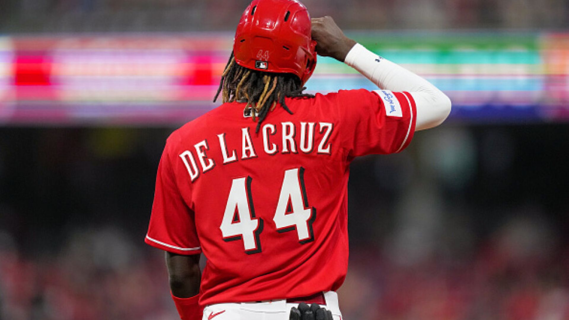 Jugador de béisbol Elly De La Cruz con su camiseta número 44, de pie en el campo de juego, mientras hace un gesto de saludo.