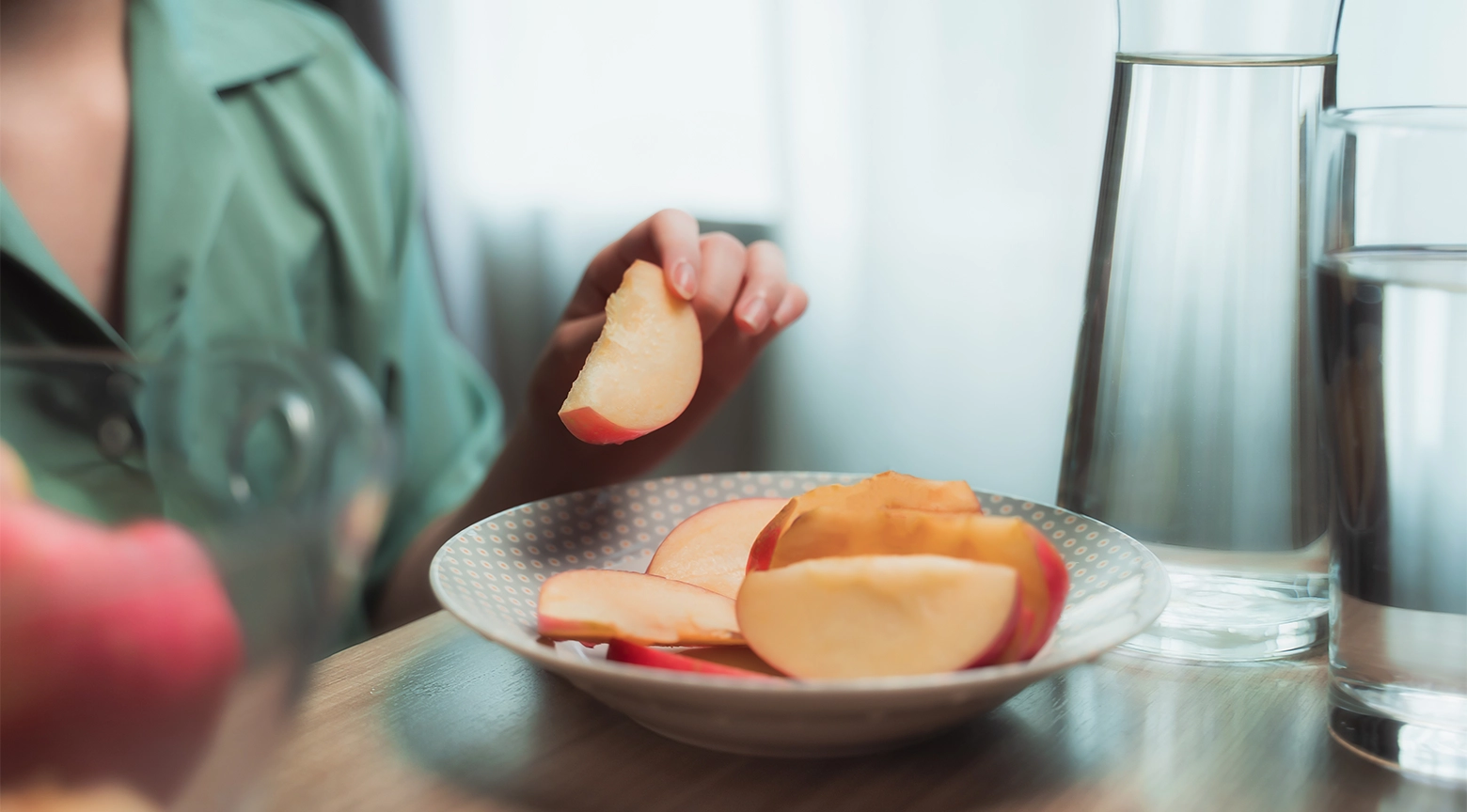 Persona comiendo una rodaja de manzana con un plato de manzana en la mesa y vasos de agua al fondo.