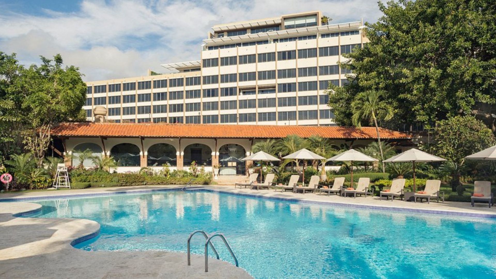 Piscina exterior de hotel con tumbonas y sombrillas frente a edificio de estilo clásico.