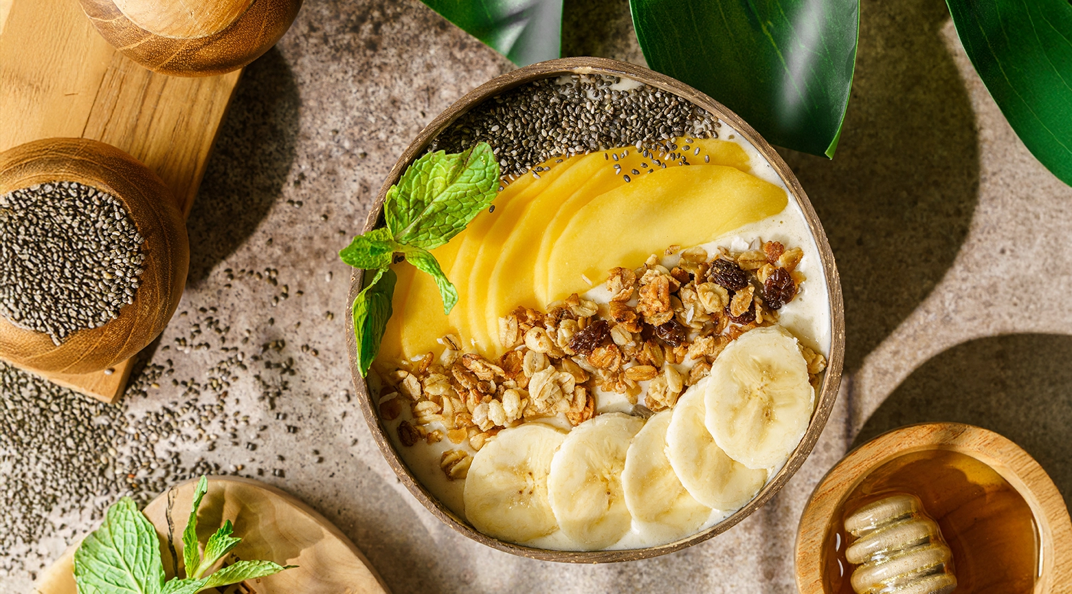 Tazón de desayuno con yogur, rodajas de plátano, granola, semillas de chía, rodajas de mango y una ramita de menta fresca.