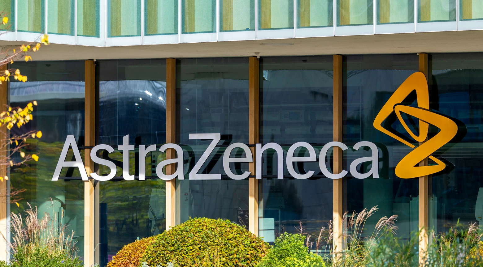 Logo de AstraZeneca en la fachada de su edificio corporativo, con reflejos de vidrio y vegetación frente a la entrada.