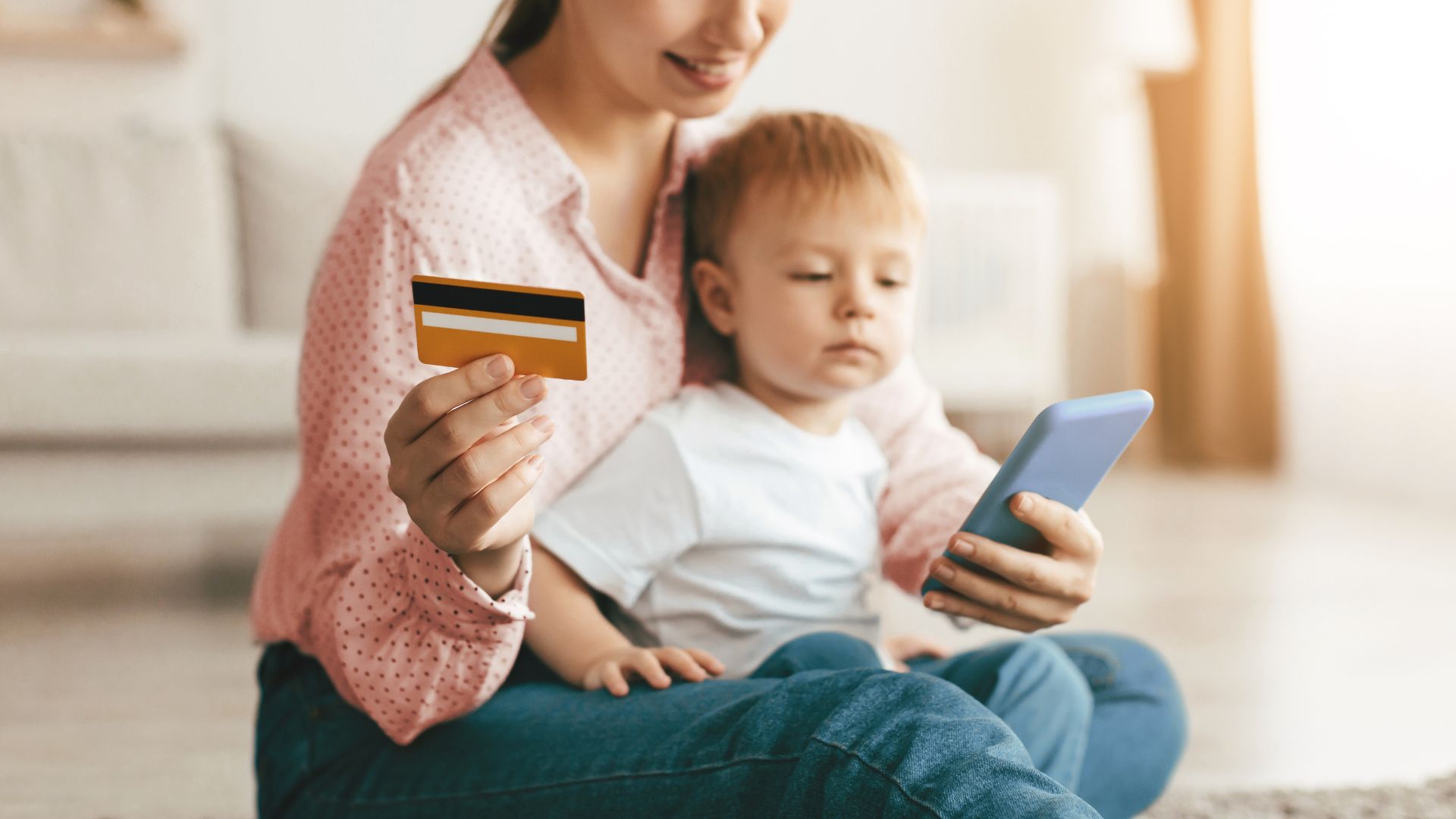 Mujer con niño pequeño revisando su teléfono mientras sostiene una tarjeta de débito, indicando una transacción o consulta de beneficio social desde casa.