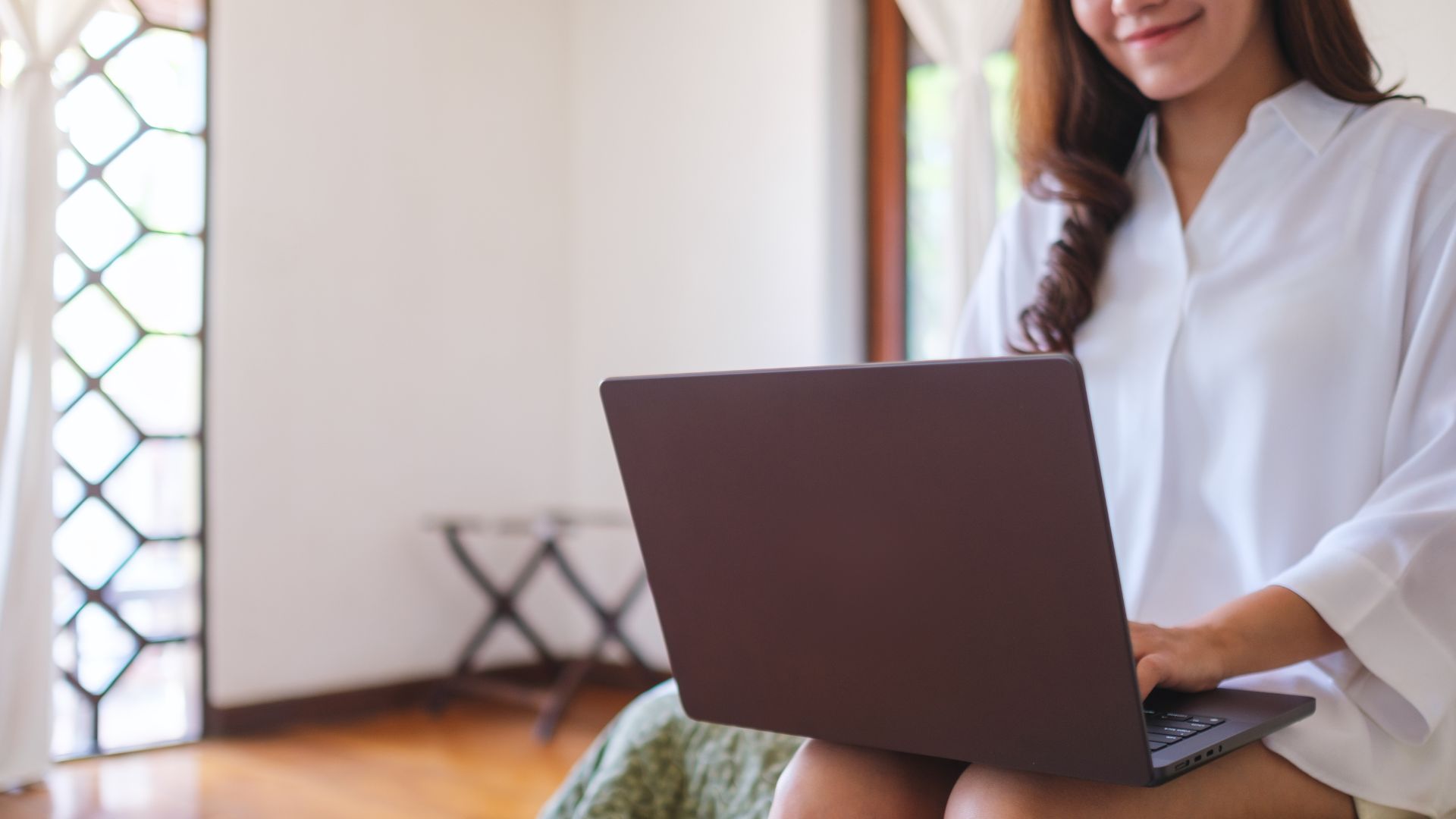 Mujer usando una computadora portátil mientras se encuentra sentada en un sofá, mostrando una expresión concentrada y sonriente, en un entorno interior iluminado naturalmente.