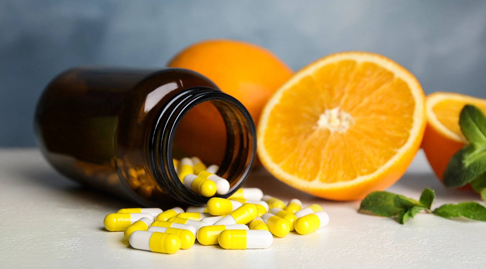 Frasco de vitaminas abierto junto a cápsulas y naranjas frescas sobre una mesa.