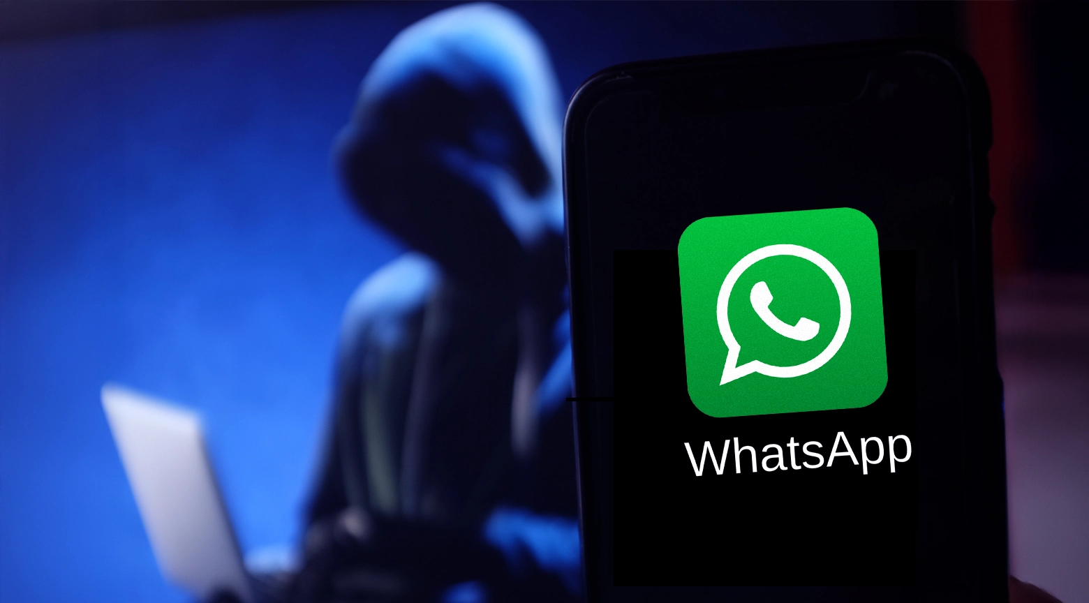 Persona con capucha utilizando una computadora portátil, mientras en primer plano se muestra el ícono de WhatsApp en un teléfono móvil, representando una cuenta hackeada.