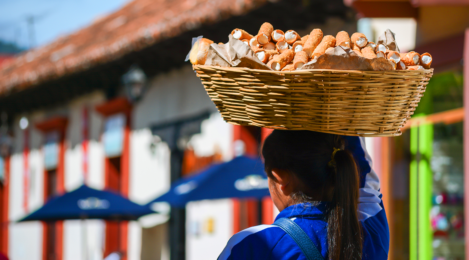 Mujer transportando una canasta grande de dulces tradicionales sobre su cabeza en un entorno urbano colorido, con edificios y sombrillas desenfocados al fondo.