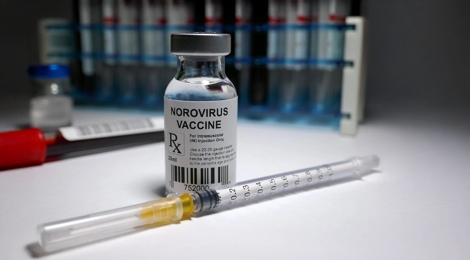 Frasco etiquetado como vacuna contra el norovirus junto a una jeringa sobre una mesa de laboratorio.