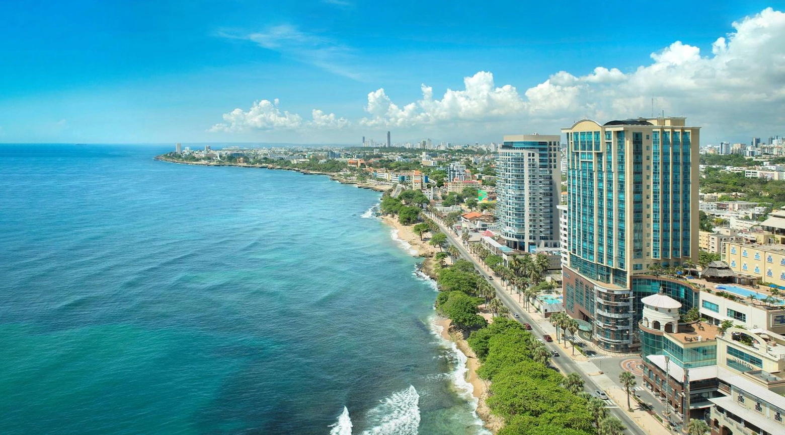 Vista aérea del litoral de Santo Domingo con edificios modernos, avenida costera y el mar Caribe bajo un cielo despejado.