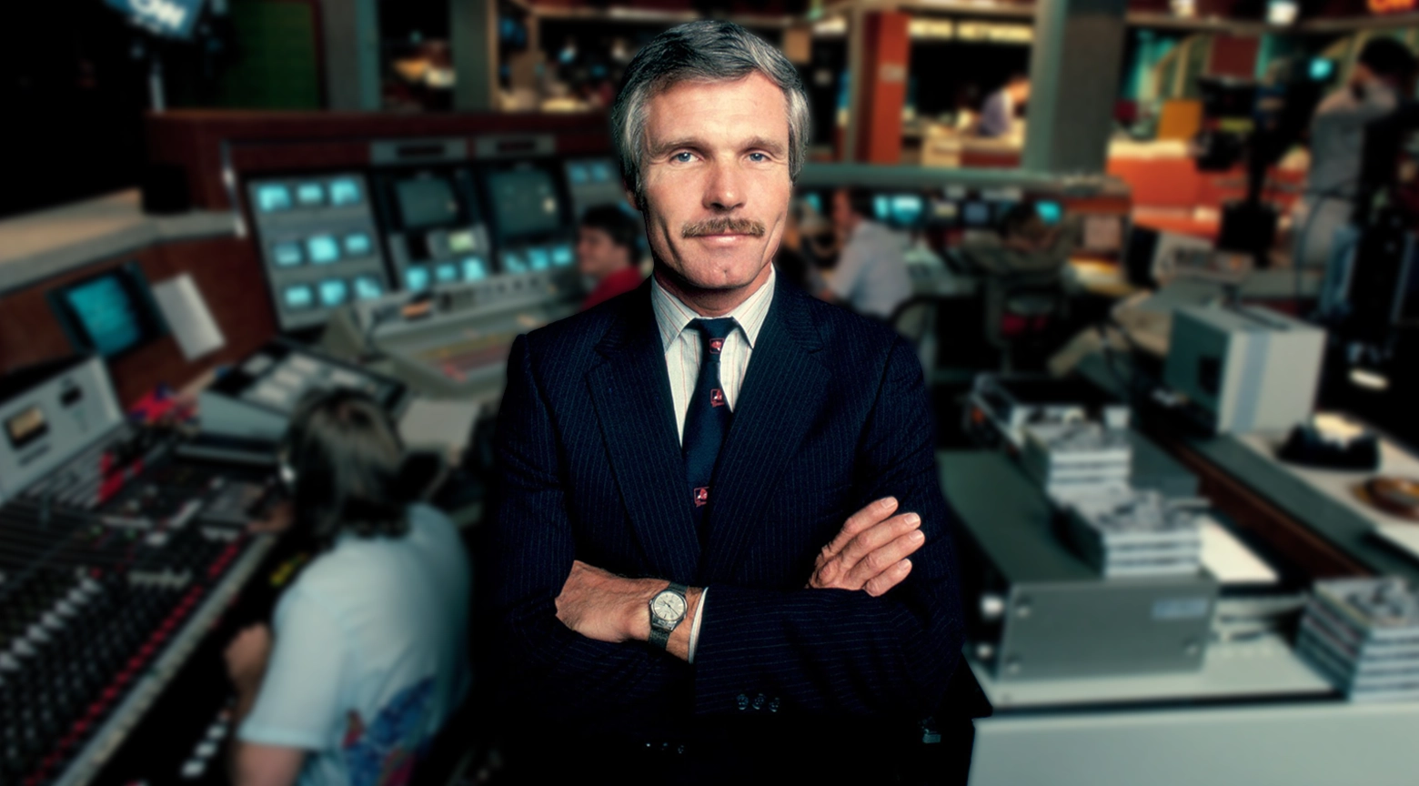 Ted Turner posa con los brazos cruzados dentro de una sala de redacción y producción televisiva.