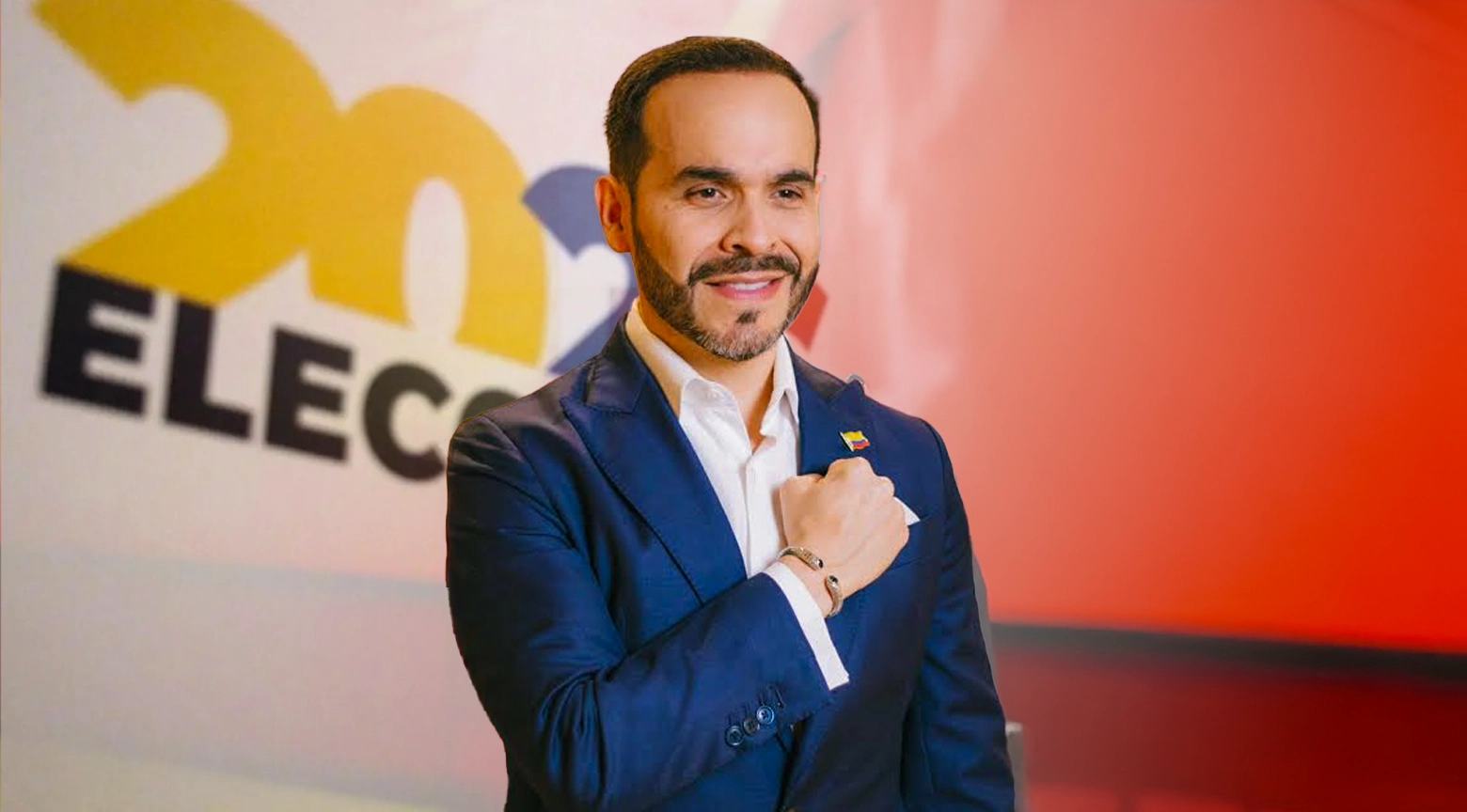 Plano medio de frente del candidato presidencial Abelardo de la Espriella con barba recortada fina vistiendo un traje formal azul oscuro sobre una camisa blanca de cuello abierto sin corbata con un pequeño pin de la bandera de Colombia en la solapa izquierda de su chaqueta mientras sonríe levemente y posa con el puño cerrado de su mano derecha apoyado sobre el pecho luciendo una pulsera metálica en su muñeca todo delante de un fondo publicitario institucional dividido horizontalmente en una mitad superior blanca con letras negras y otra mitad inferior de color rojo sólido.