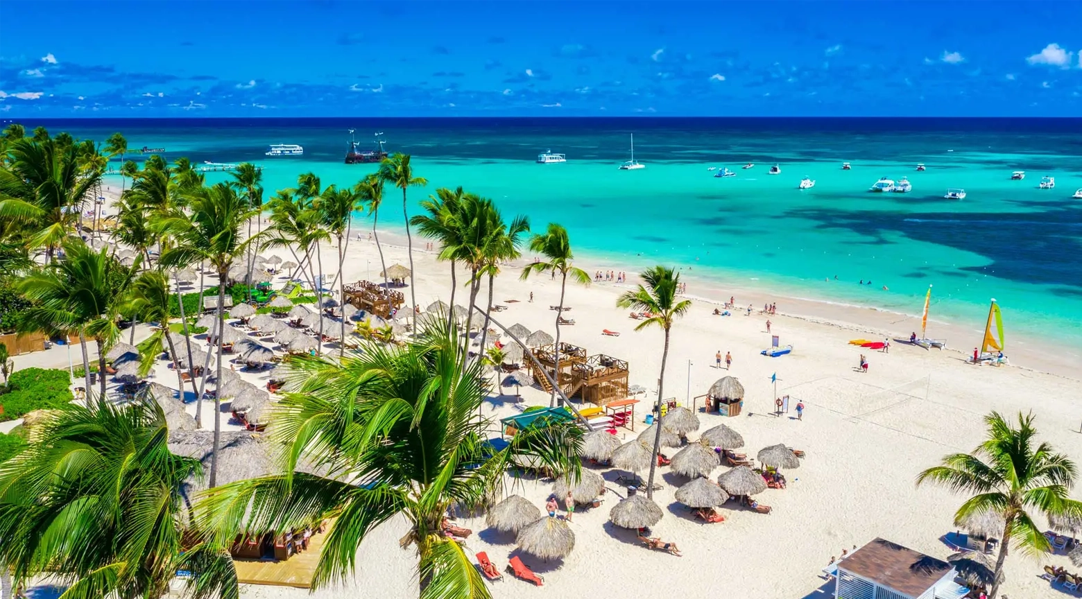 Playa tropical en República Dominicana con arena blanca, palmeras, sombrillas de paja, turistas y actividades acuáticas en el mar turquesa.