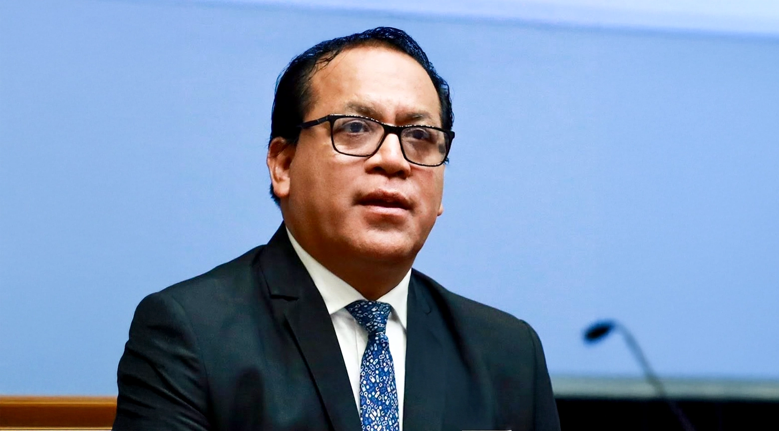 Retrato en primer plano medio del candidato izquierdista Roberto Sánchez, con gafas, traje oscuro, camisa blanca y corbata azul estampada, mirando hacia la derecha con expresión seria mientras habla durante una conferencia de prensa con un fondo azul claro uniforme.