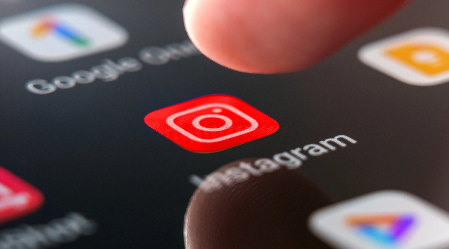 Primer plano extremo de la pantalla encendida de un teléfono inteligente donde destaca en el centro el icono cuadrado de la aplicación de Instagram con su característico diseño de cámara en color blanco sobre un fondo degradado de tonos rojos naranjas y morados mientras el dedo de una persona aparece desenfocado en la parte superior a punto de tocar la pantalla rodeado de otros accesos directos borrosos como la suite corporativa de Google.