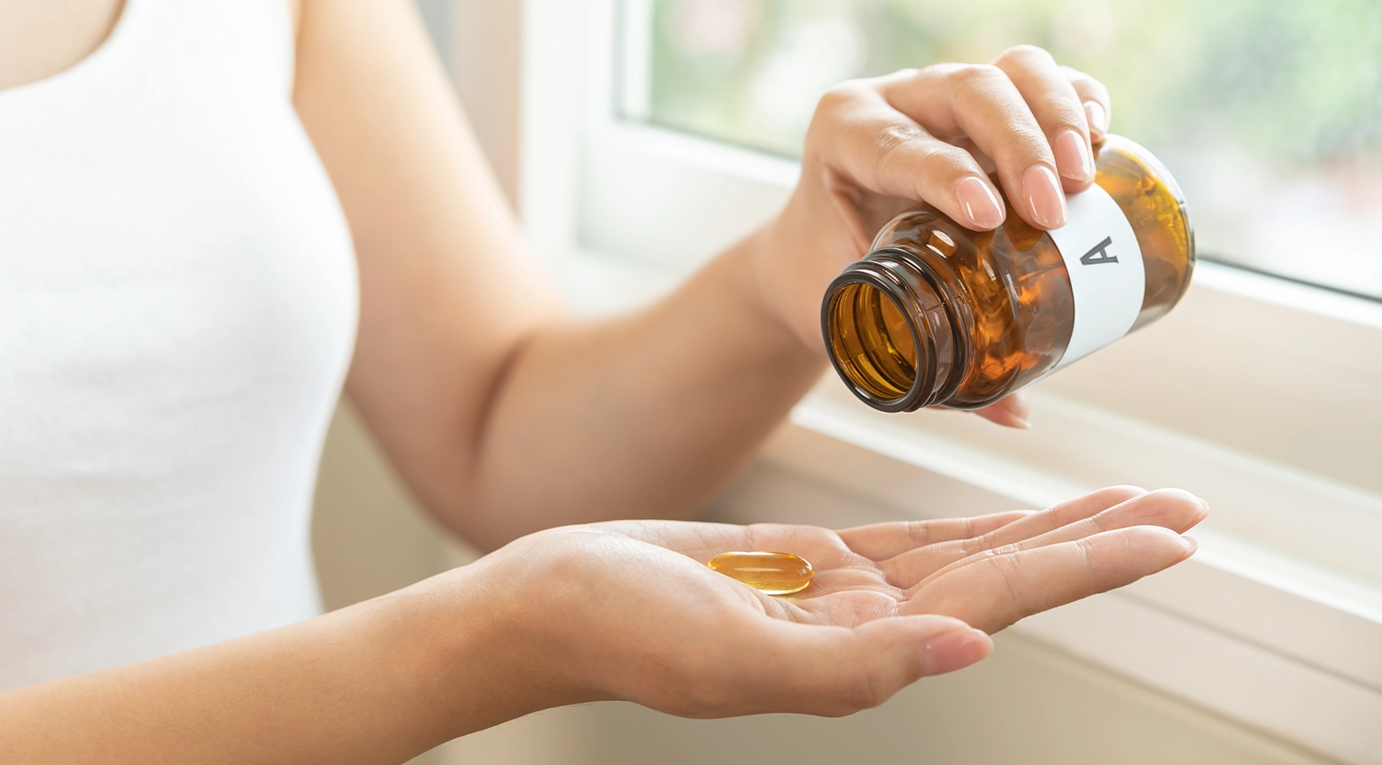 Persona sosteniendo un frasco de suplementos vitamínicos mientras deja caer una cápsula en la palma de su otra mano cerca de una ventana con luz natural.
