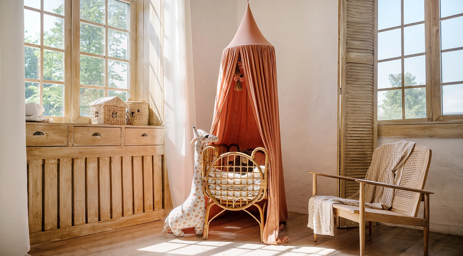 Habitación infantil de estilo rústico iluminada por la luz del sol con una cuna de mimbre vacía bajo un dosel rosado y una jirafa de peluche al lado de un ventanal de madera.