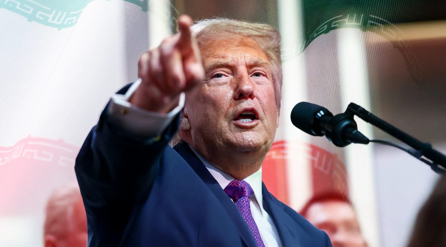 Plano medio de frente de Donald Trump vistiendo un traje azul oscuro con camisa blanca y una corbata de tonos morados con patrones geométricos mientras habla de frente ante un micrófono negro doble fijado en un soporte articulado y extiende su brazo derecho hacia el frente apuntando directamente con el dedo índice en un gesto enfático teniendo de fondo una imagen superpuesta y difuminada que combina la bandera de Irán con sus colores verde blanco y rojo junto al escudo nacional central.