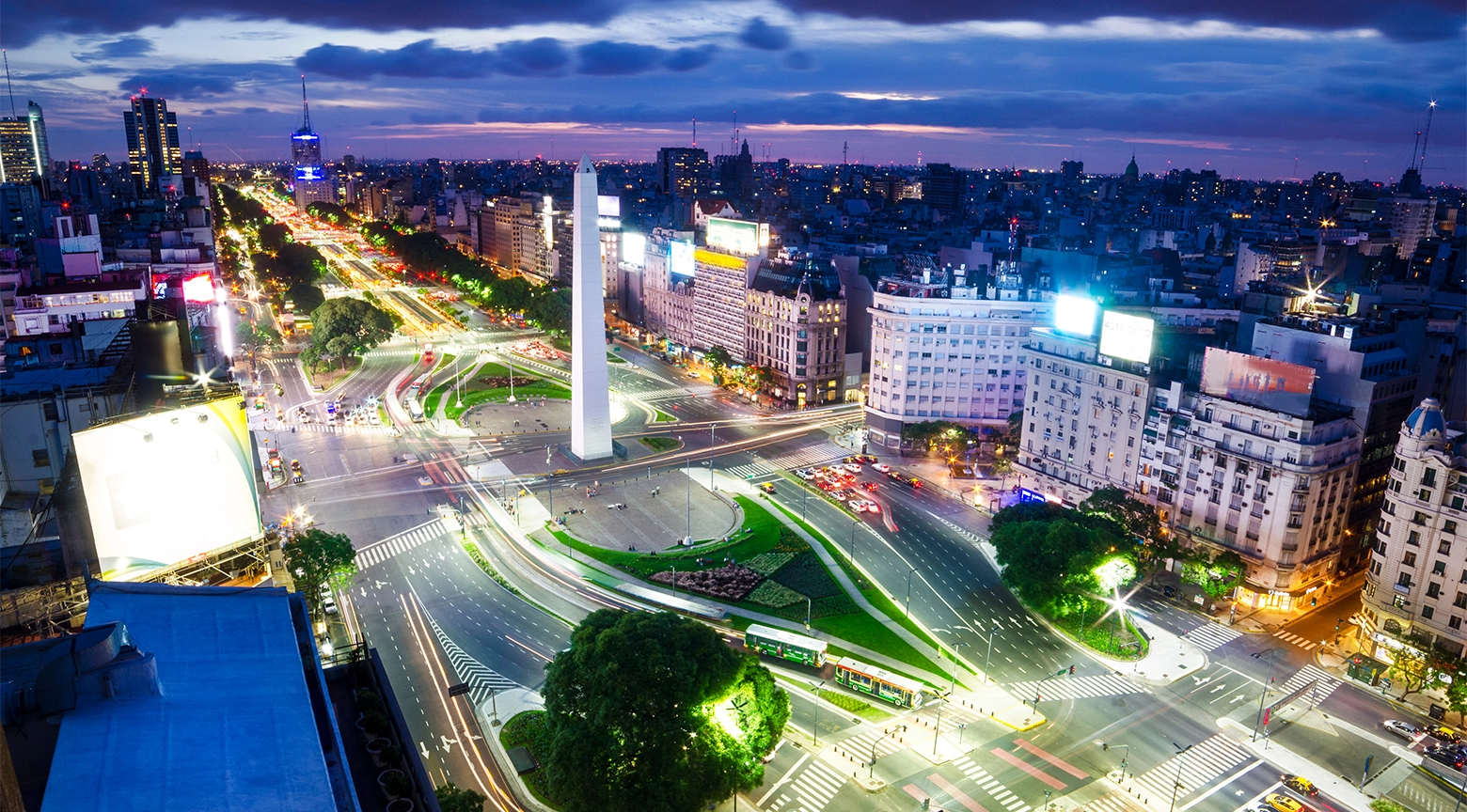 Vista panorámica nocturna de Buenos Aires con el Obelisco iluminado, avenidas con tráfico y edificios urbanos en el centro de la ciudad.
