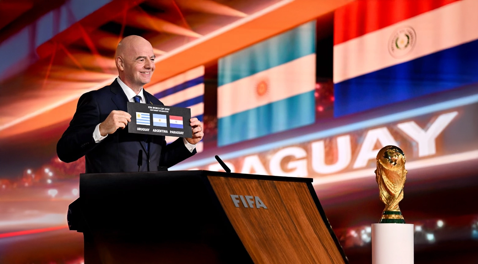 Gianni Infantino sosteniendo una tarjeta con las banderas de Uruguay, Argentina y Paraguay durante el anuncio de las sedes de la Copa Mundial 2030, con la copa del mundo a su lado en el podio.