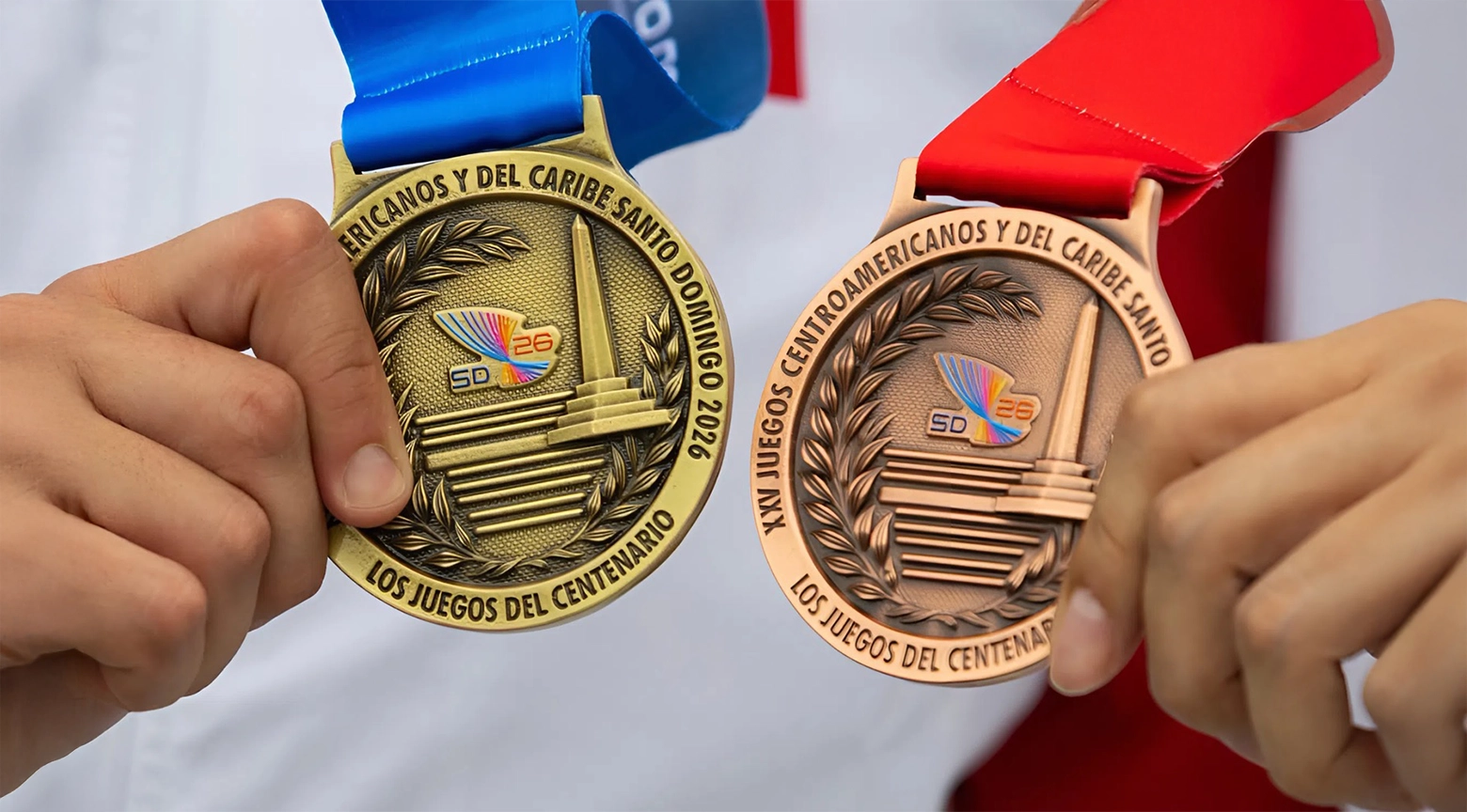 Dos manos sostienen las medallas de oro y bronce de los XXV Juegos Centroamericanos y del Caribe Santo Domingo 2026.