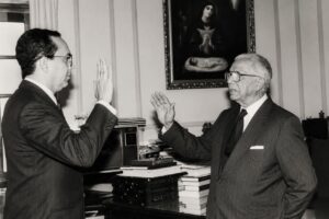 Fotografía de archivo en blanco y negro de Héctor Valdez Albizu durante una ceremonia institucional, con ambos participantes levantando la mano derecha.