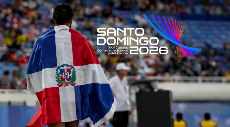 Atleta con la bandera de República Dominicana sobre los hombros en un estadio, con el logotipo de los Juegos Centroamericanos y del Caribe Santo Domingo 2026 al fondo.