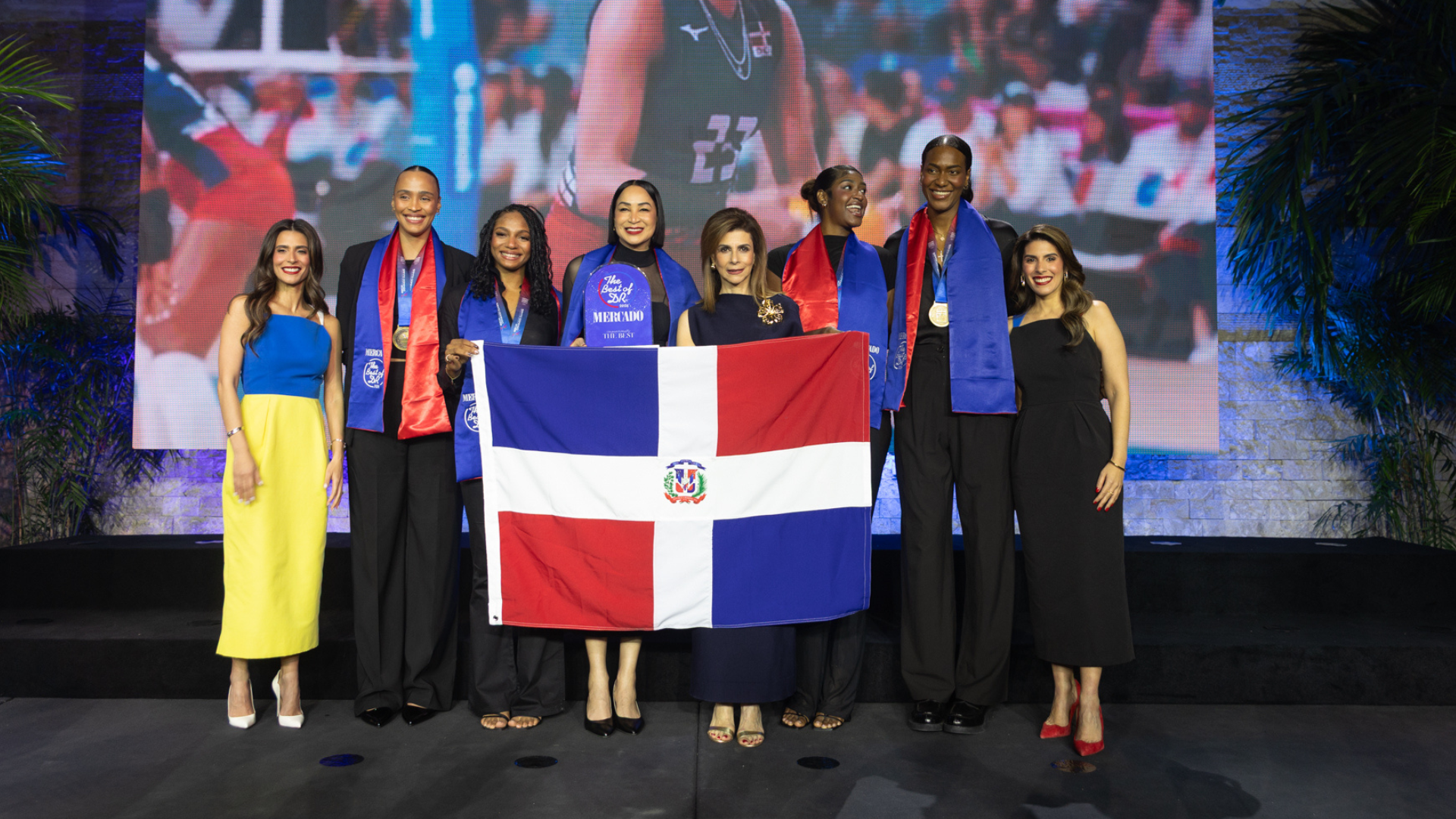 Grupo de personas en un escenario sostiene una bandera de República Dominicana; detrás se proyecta una imagen de una jugadora de voleibol durante un partido.