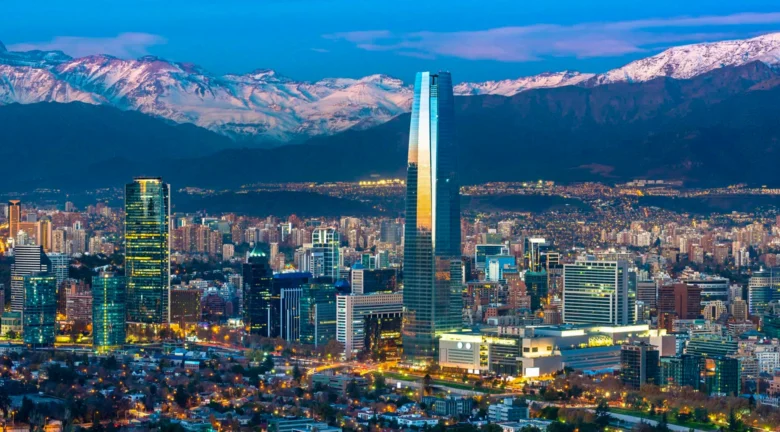 Vista panorámica de una ciudad moderna al atardecer, con un rascacielos de cristal destacado en primer plano y montañas nevadas al fondo.