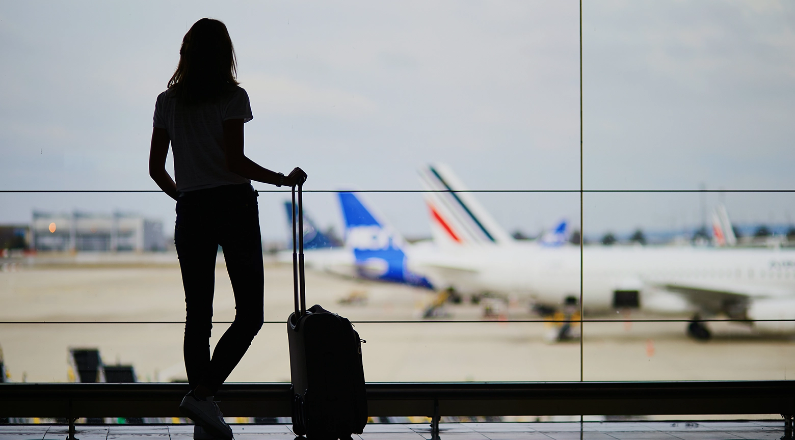 Silueta de una mujer con una maleta de viaje frente a una ventana de aeropuerto, con aviones estacionados en la pista al fondo.