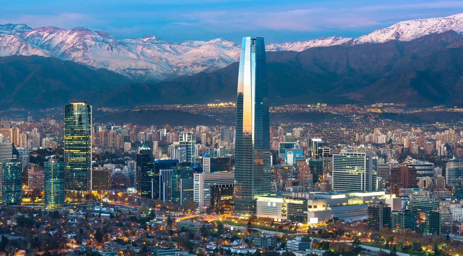 Vista panorámica de una ciudad latinoamericana moderna con edificios altos, una torre de oficinas destacada y montañas nevadas al fondo, representando desarrollo urbano y cambios demográficos.