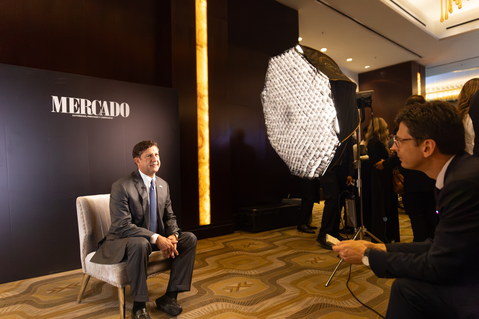 Entrevista profesional en un evento corporativo: un ejecutivo con traje sentado frente a un equipo de grabación, con un fondo de la marca Mercado y personal realizando una sesión audiovisual.