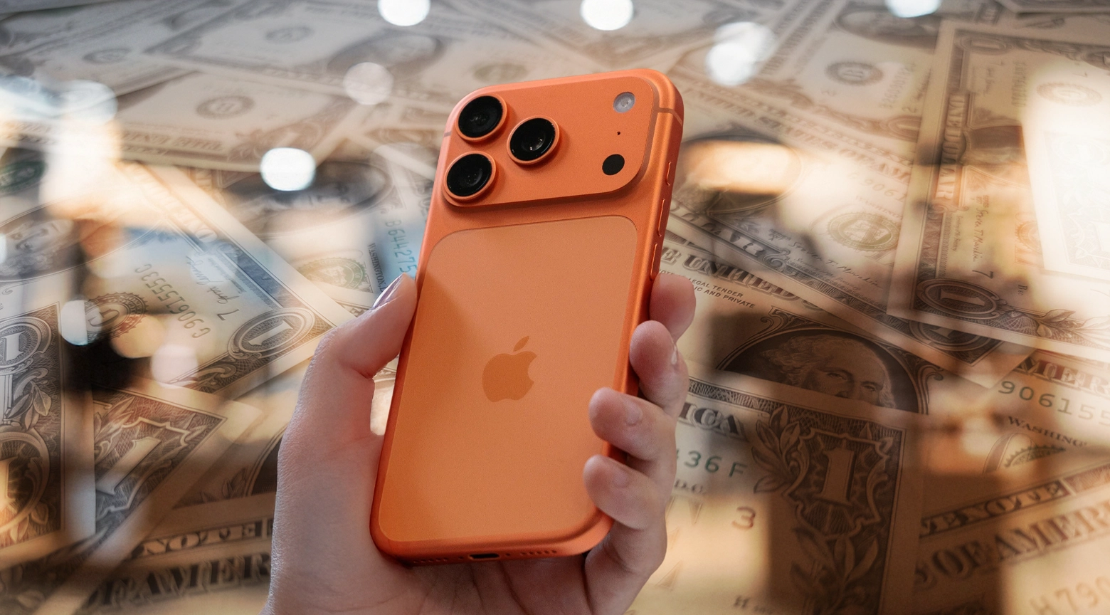 Una mano sostiene un iPhone de color naranja sobre un fondo compuesto por billetes de dólares estadounidenses.