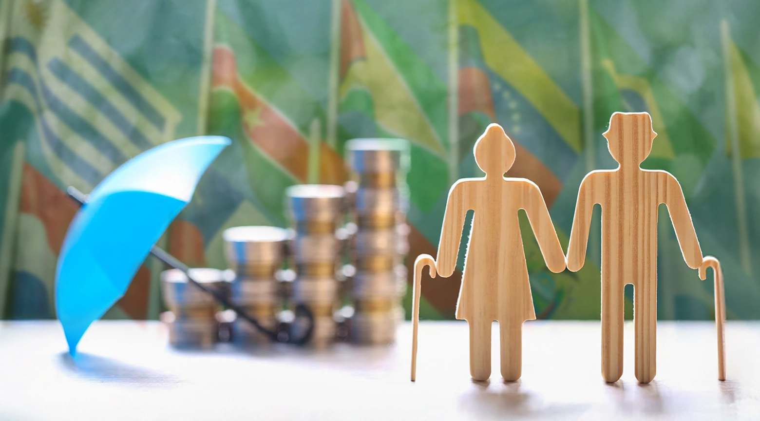 Figuras de madera que representan a personas mayores junto a monedas apiladas y un paraguas azul, con banderas de países de América Latina desenfocadas al fondo.