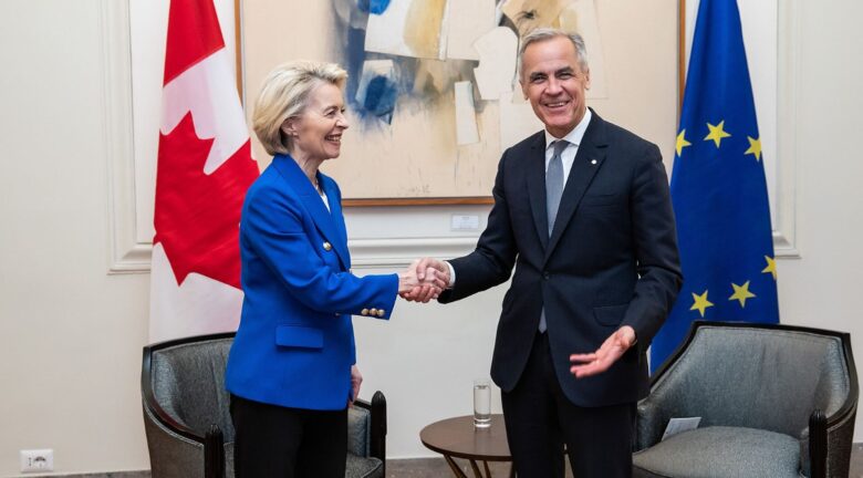 Ursula von der Leyen y Mark Carney estrechan sus manos durante un encuentro institucional, con las banderas de Canadá y la Unión Europea al fondo.