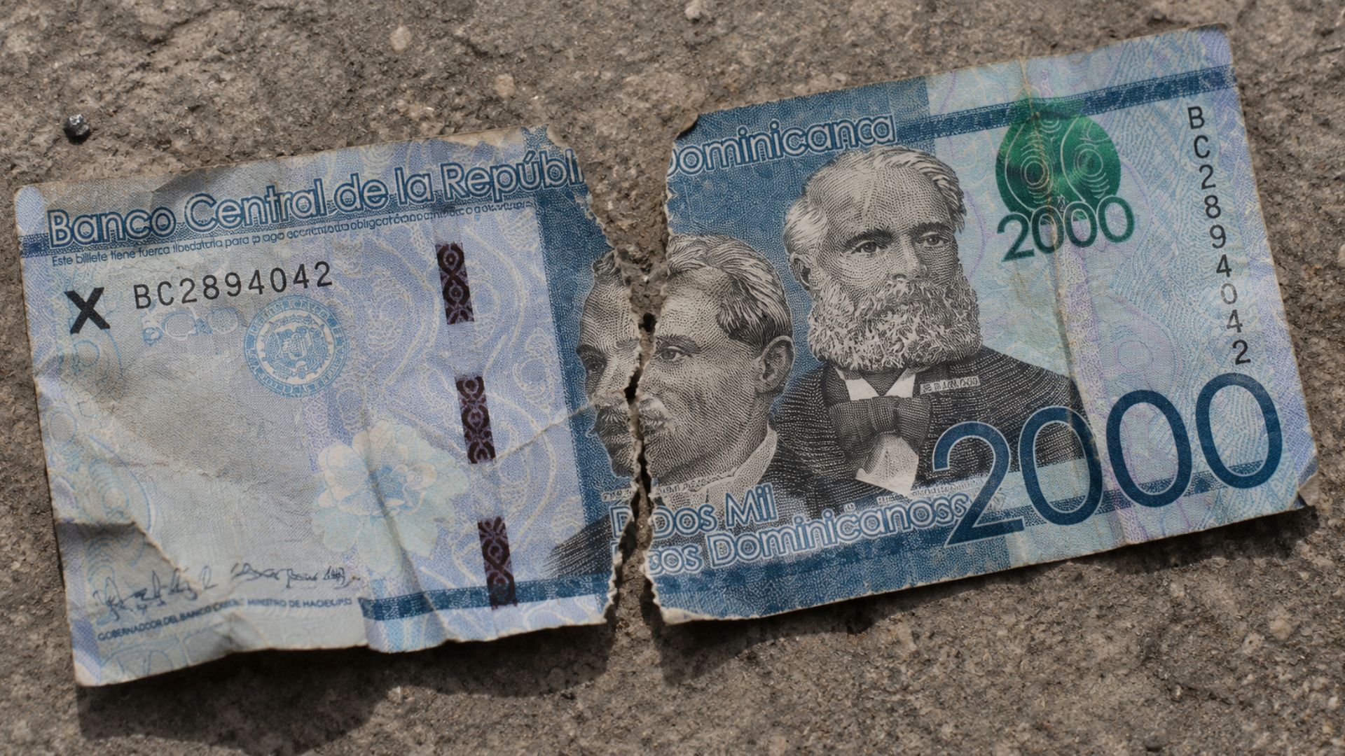 Billete dominicano de 2,000 pesos roto en dos partes sobre una superficie de concreto.