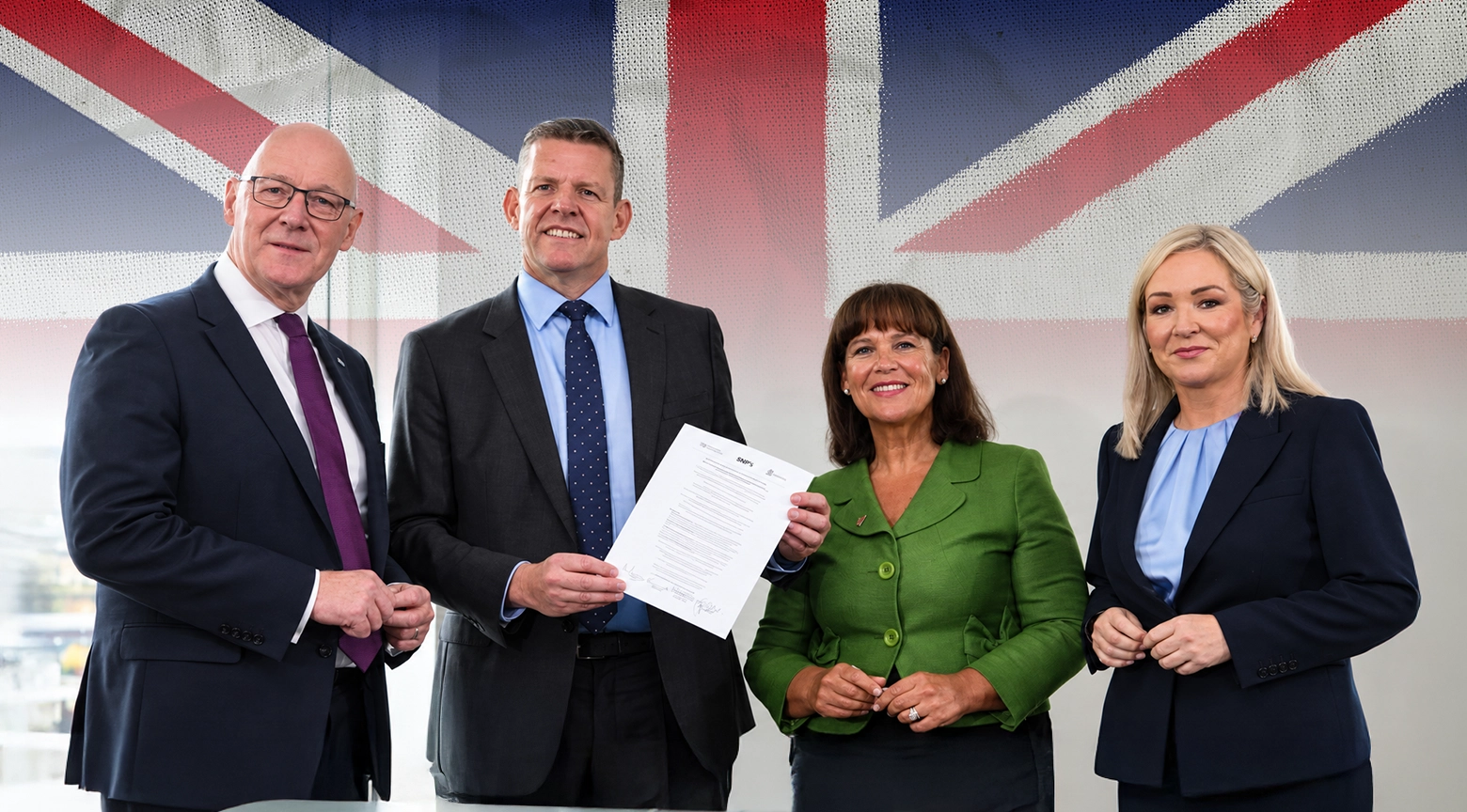 Cuatro políticos sonrientes en traje formal posan juntos sosteniendo un documento frente a la bandera del Reino Unido (Union Jack).