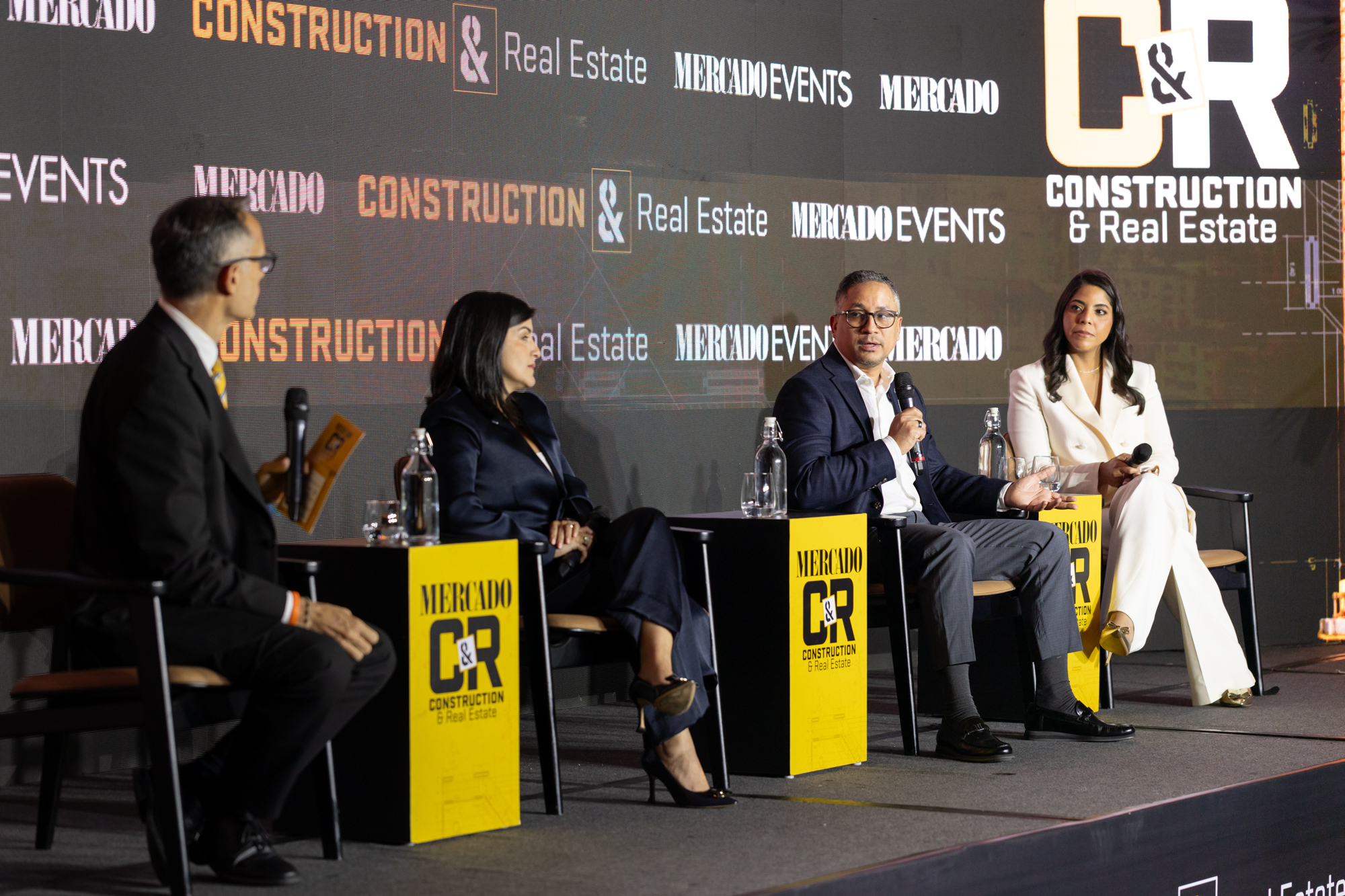 Panel de conversación sobre construcción y bienes raíces, con cuatro participantes sentados en el escenario y el logotipo de Mercado C&R Construction & Real Estate proyectado al fondo.