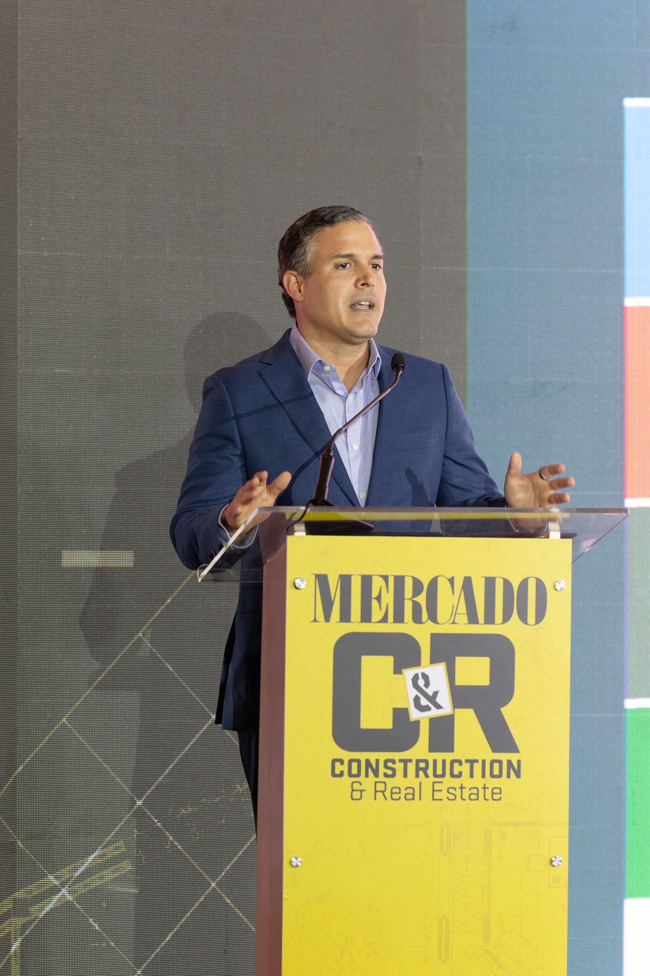 Hombre con traje azul y camisa clara dando un discurso detrás de un podio de Mercado C&R Construction & Real Estate, frente a una pantalla LED.