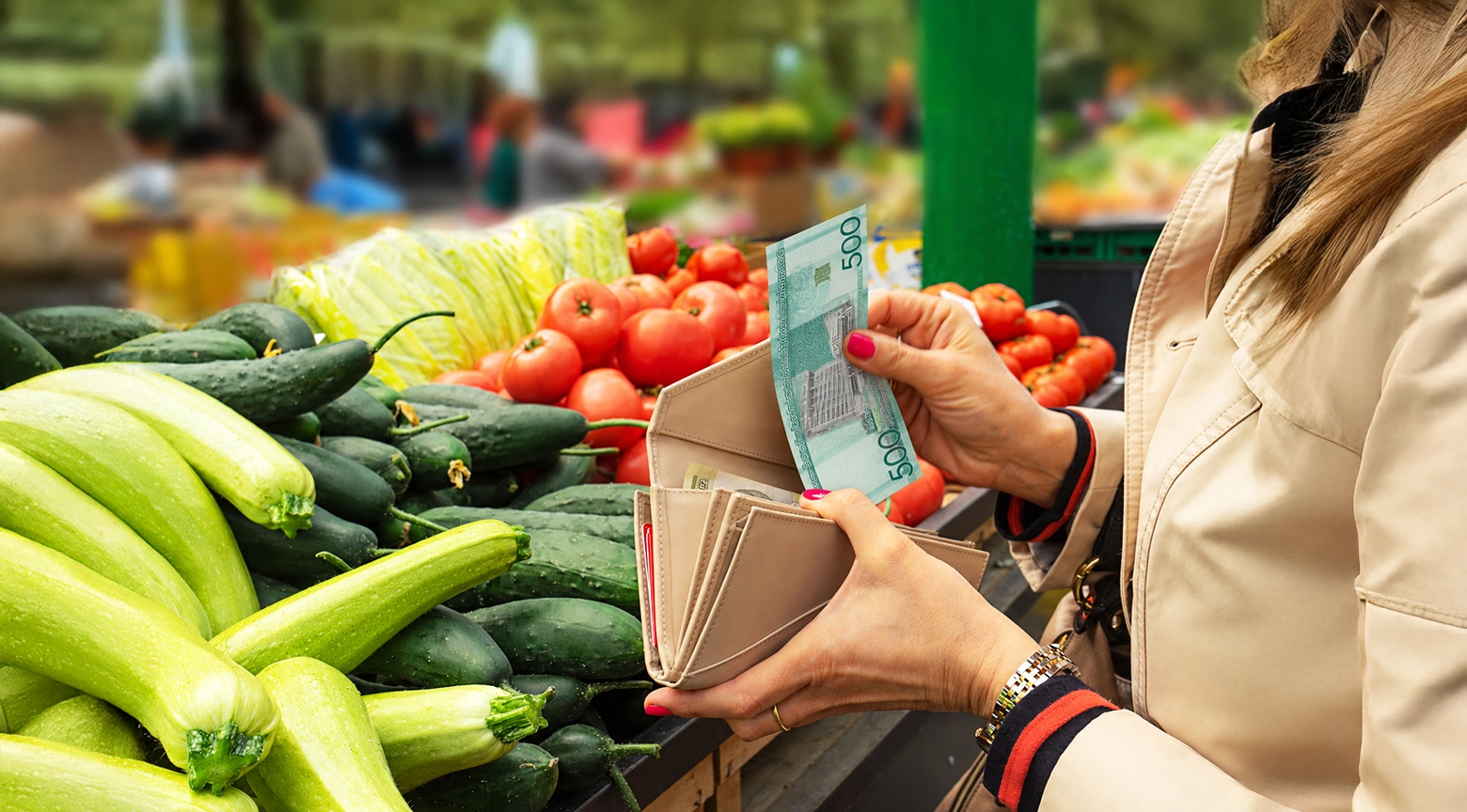 Mujer sostiene un billete de 500 pesos dominicanos y una cartera frente a un puesto de verduras con tomates, pepinos y calabacines, en una representación del costo de la canasta básica y el poder adquisitivo.
