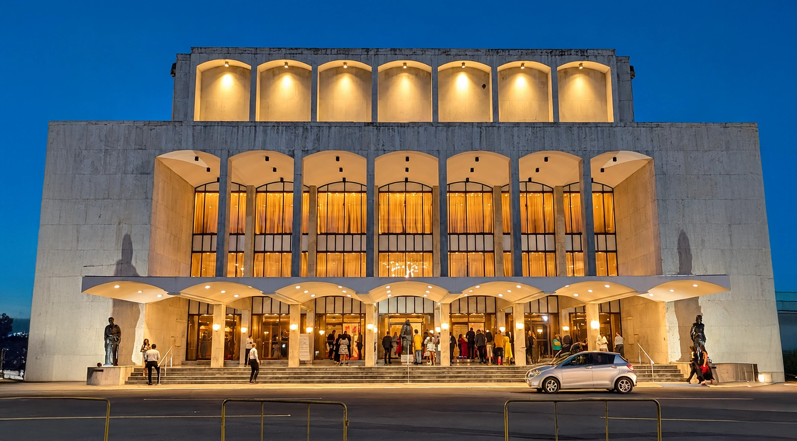Vista frontal del Teatro Nacional Eduardo Brito, en Santo Domingo, al anochecer. Su fachada de piedra clara aparece iluminada con luces cálidas, mientras varios visitantes se reúnen en las escalinatas y frente a la entrada principal.