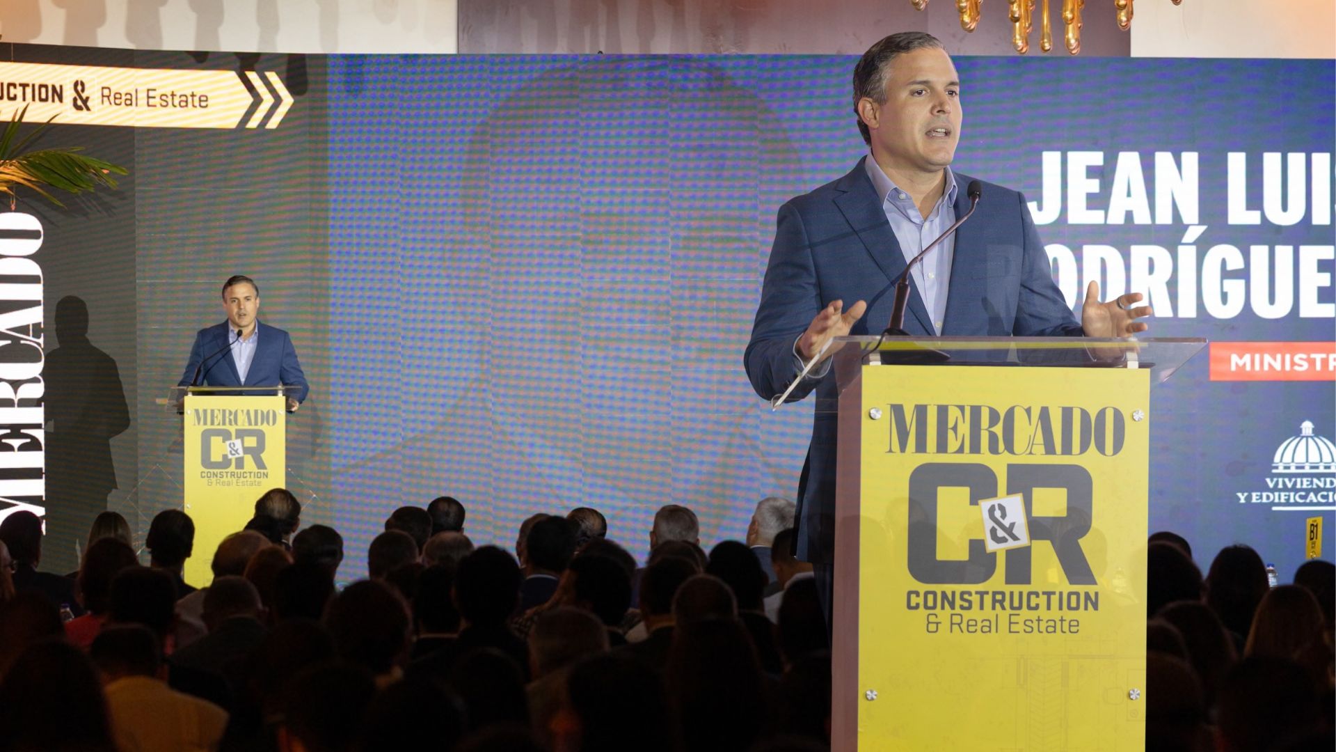 Hombre dando un discurso desde un podio de Mercado C&R Construction & Real Estate, ante un público, mientras una pantalla gigante muestra su imagen y datos de su participación.
