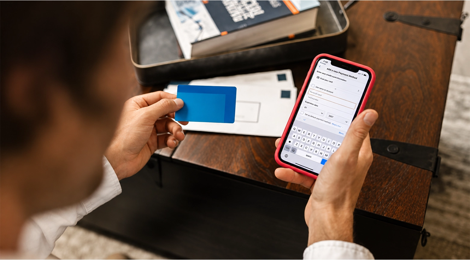 Una persona sostiene una tarjeta bancaria azul y un teléfono inteligente con una funda roja. En la pantalla aparece un formulario para introducir los datos de una tarjeta como método de pago, mientras al fondo se observan documentos y un libro sobre una mesa de madera.