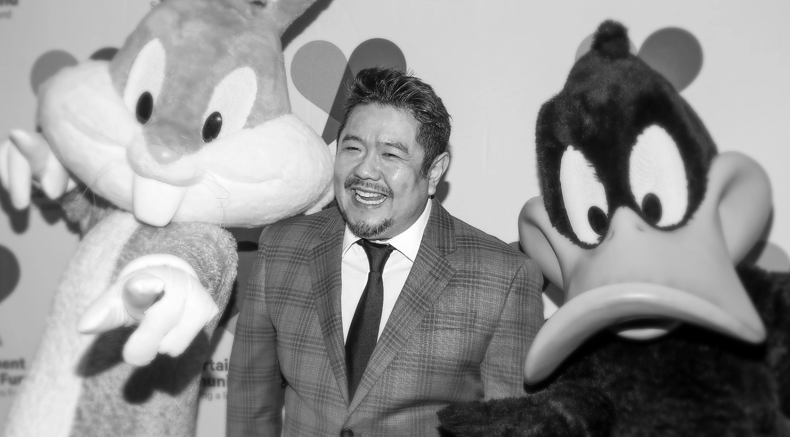 Fotografía en blanco y negro del actor de voz Eric Bauza, vestido con traje y corbata, posando sonriente entre dos personajes de Looney Tunes: Bugs Bunny, a la izquierda, y el Pato Lucas, a la derecha, durante un evento público.