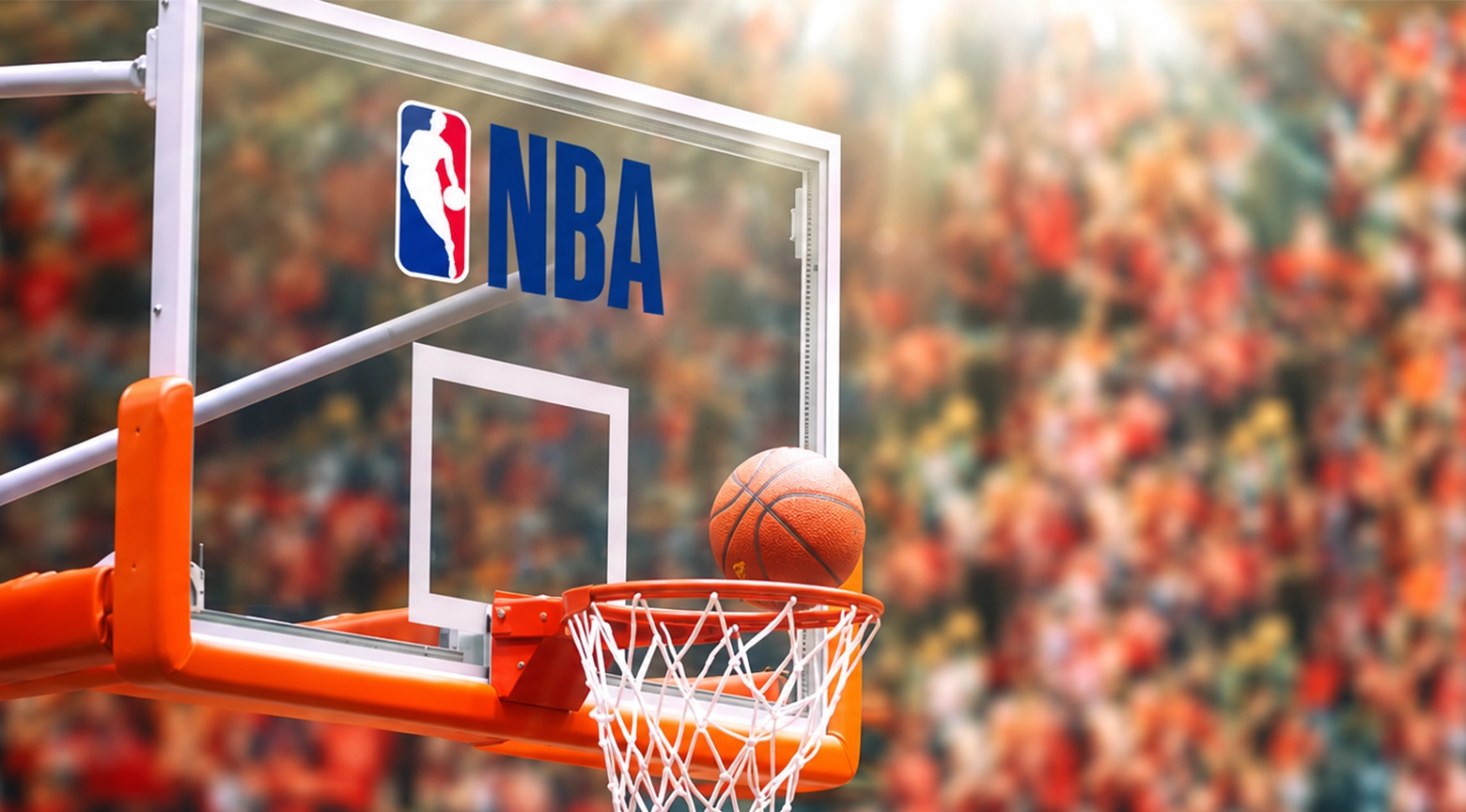 Primer plano de una canasta de baloncesto con tablero transparente y el logotipo de la NBA. Un balón naranja aparece sobre el aro, mientras al fondo se observa una multitud de aficionados desenfocados en las gradas de un estadio.