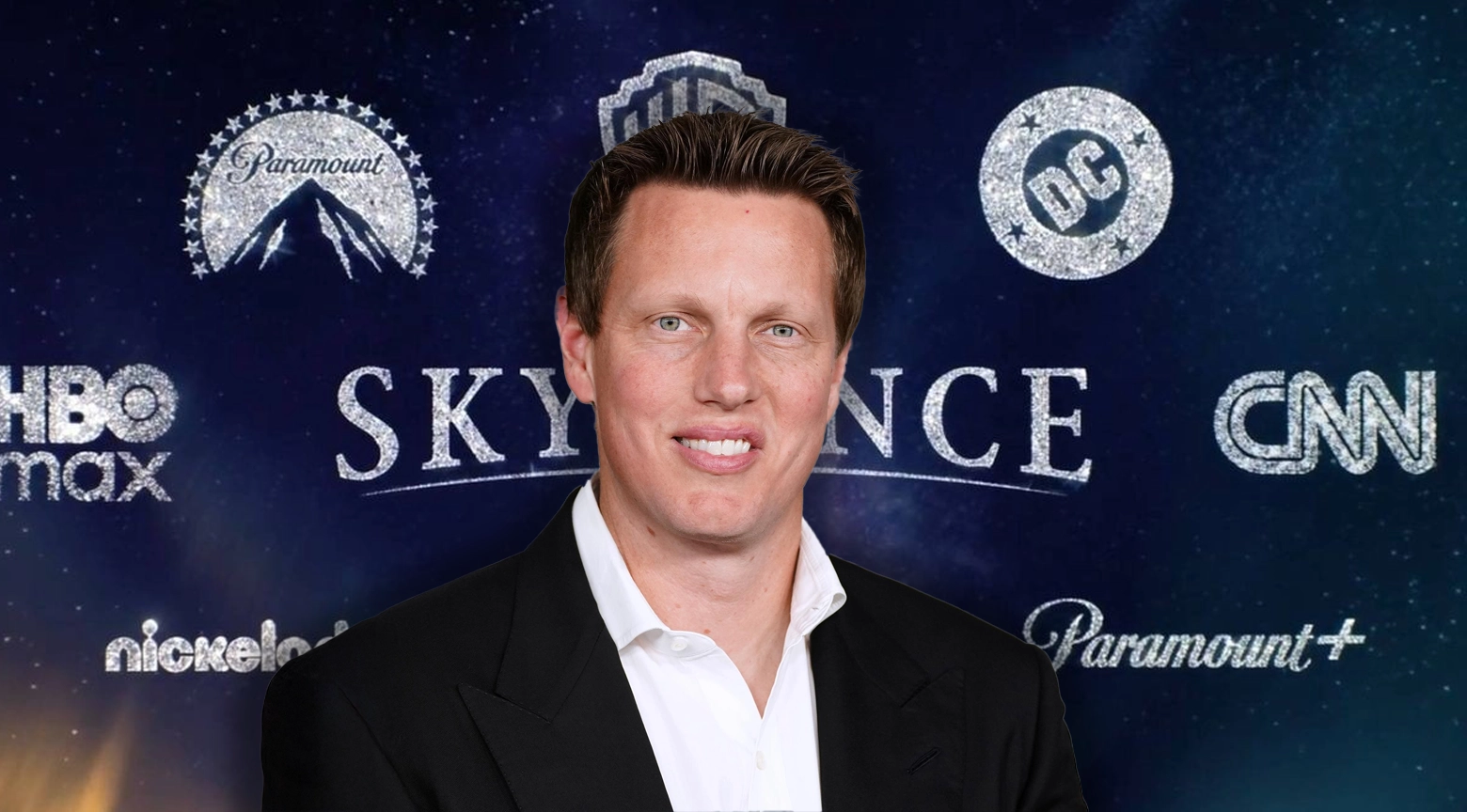 Hombre vestido con traje negro y camisa blanca frente a un fondo con los logotipos de Paramount, Warner Bros., DC, HBO Max, CNN y otras marcas de entretenimiento.