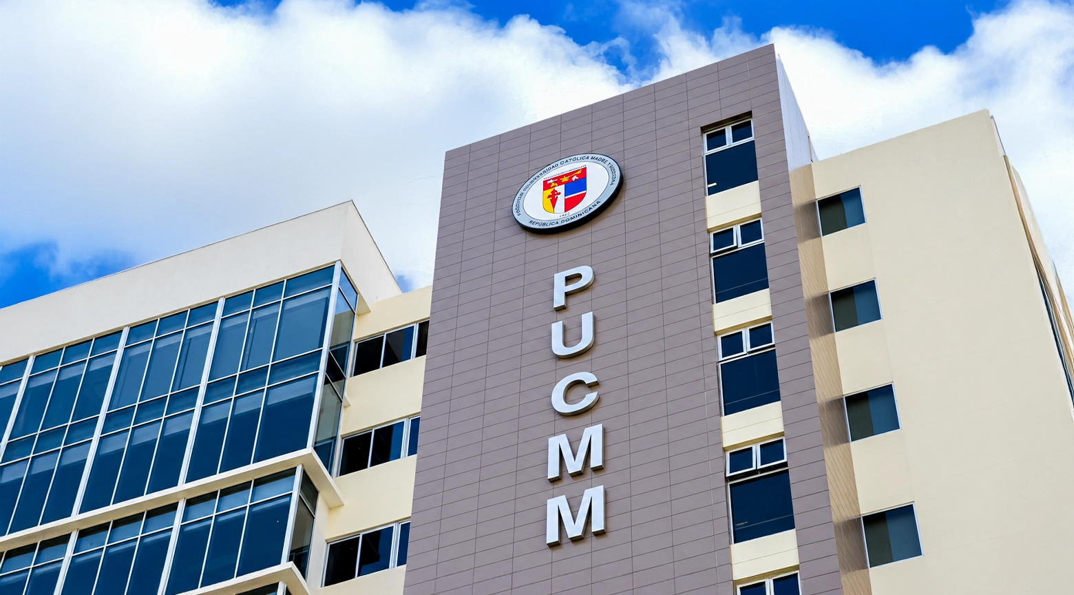 Fachada de un edificio de la Pontificia Universidad Católica Madre y Maestra (PUCMM), en República Dominicana, con el escudo institucional y las siglas PUCMM colocadas verticalmente sobre una estructura moderna de cristal y concreto.