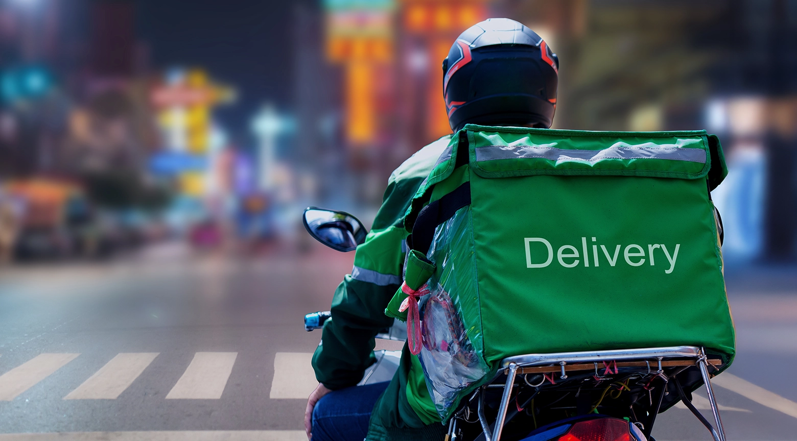 Repartidor de comida a domicilio en motocicleta, vestido con uniforme verde y casco de seguridad, transporta una mochila térmica con la palabra «Delivery» por una calle urbana.